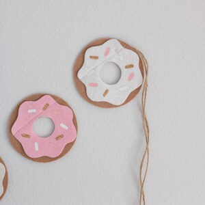 K&ouml;nnte beinhalten: Eine Girlande aus Filzdonuts mit rosa und wei&szlig;em Zuckerguss und goldenen Streuseln. Die Donuts sind mit einer Schnur aneinandergereiht.