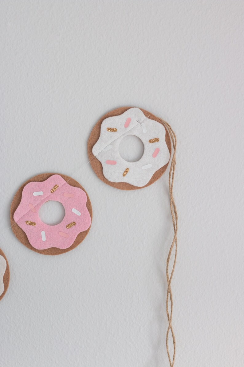 K&ouml;nnte beinhalten: Eine Girlande aus Filzdonuts mit rosa und wei&szlig;em Zuckerguss und goldenen Streuseln. Die Donuts sind mit einer Schnur aneinandergereiht.