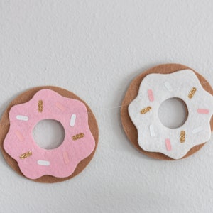 K&ouml;nnte beinhalten: Eine Girlande aus Filzdonuts mit rosa und wei&szlig;em Zuckerguss und goldenen Streuseln. Die Donuts sind mit wei&szlig;em Garn zusammengef&uuml;gt.