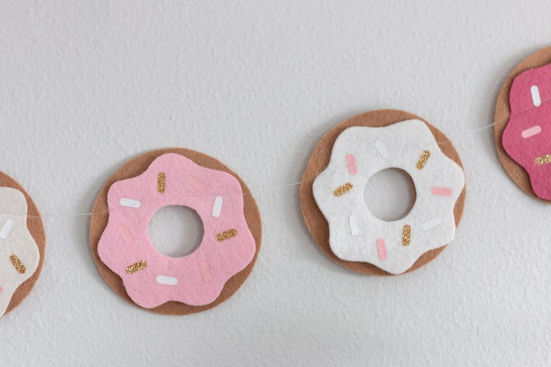 K&ouml;nnte beinhalten: Eine Girlande aus Filzdonuts mit rosa und wei&szlig;em Zuckerguss und goldenen Streuseln. Die Donuts sind mit wei&szlig;em Garn zusammengef&uuml;gt.
