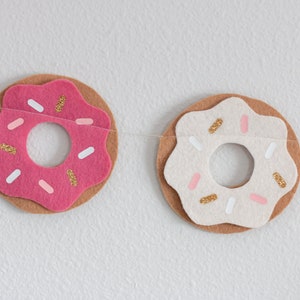 K&ouml;nnte beinhalten: Eine Girlande aus rosa und wei&szlig;en Filzdonuts mit goldenen Streuseln. Die Donuts sind mit einem durchsichtigen Faden aneinandergereiht.