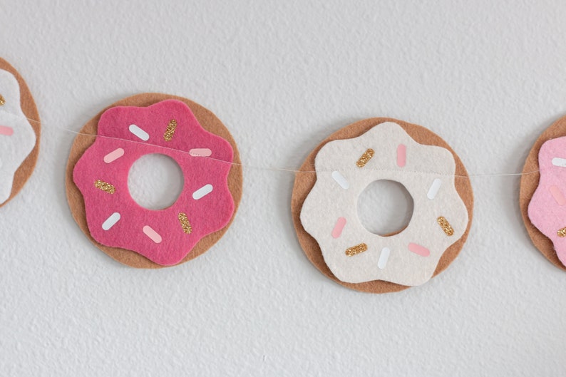 K&ouml;nnte beinhalten: Eine Girlande aus rosa und wei&szlig;en Filzdonuts mit goldenen Streuseln. Die Donuts sind mit einem durchsichtigen Faden aneinandergereiht.