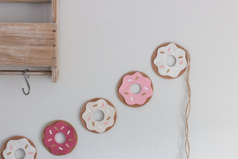 K&ouml;nnte beinhalten: Eine Girlande aus Filzdonuts mit rosa und wei&szlig;em Zuckerguss und goldenen Streuseln. Die Donuts sind mit einer Schnur aneinandergereiht.