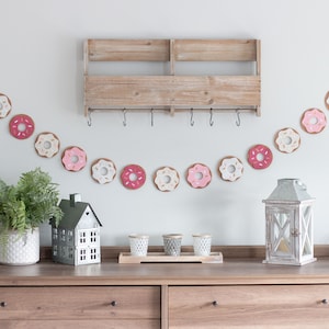 K&ouml;nnte beinhalten: Ein Holzregal mit Haken h&auml;ngt an einer wei&szlig;en Wand. Unter dem Regal h&auml;ngt eine Girlande aus donutf&ouml;rmigen Ornamenten in Rosa, Wei&szlig; und Braun. Eine kleine hausf&ouml;rmige Dekoration, ein Topf mit Farn und eine Laterne stehen auf einer Kommode aus Holz mit zwei Schubladen.