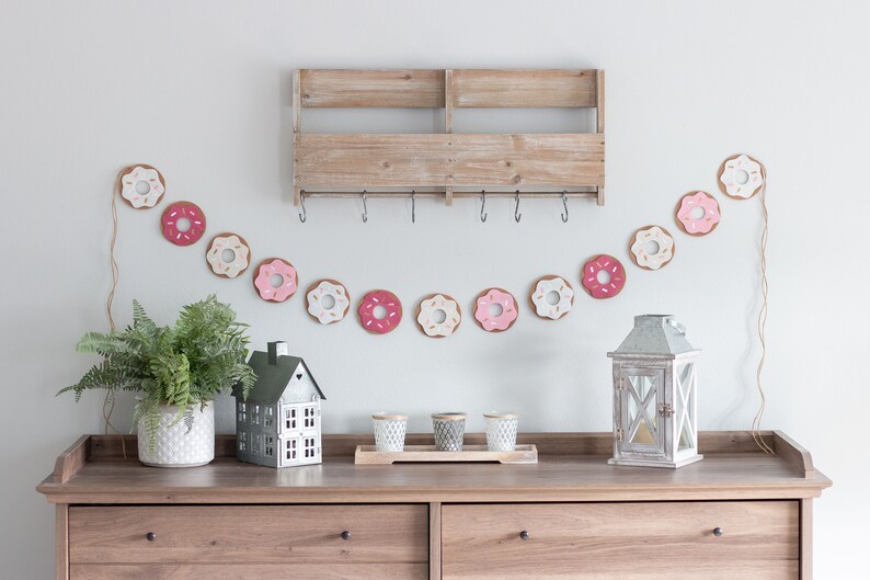K&ouml;nnte beinhalten: Ein Holzregal mit Haken h&auml;ngt an einer wei&szlig;en Wand. Unter dem Regal h&auml;ngt eine Girlande aus donutf&ouml;rmigen Ornamenten in Rosa, Wei&szlig; und Braun. Eine kleine hausf&ouml;rmige Dekoration, ein Topf mit Farn und eine Laterne stehen auf einer Kommode aus Holz mit zwei Schubladen.