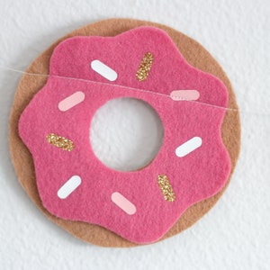 K&ouml;nnte beinhalten: Ein rosafarbener Filz-Donut mit wei&szlig;en und rosa Streuseln und goldenem Glitzer. Der Donut ist Teil einer Filz-Girlande.