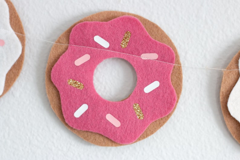 K&ouml;nnte beinhalten: Ein rosafarbener Filz-Donut mit wei&szlig;en und rosa Streuseln und goldenem Glitzer. Der Donut ist Teil einer Filz-Girlande.