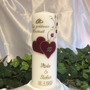 Könnte beinhalten: Weiße Kerze mit goldener Beschriftung und zwei burgunderfarbenen Herzen. Die Kerze ist mit goldenen Ranken und burgunderfarbenen Blüten verziert. Der Text auf der Kerze lautet "Zur goldenen Hochzeit", "Viola & Stefan" und "18.3.1969". Die Zahl "50" ist in eines der Herzen geschrieben.