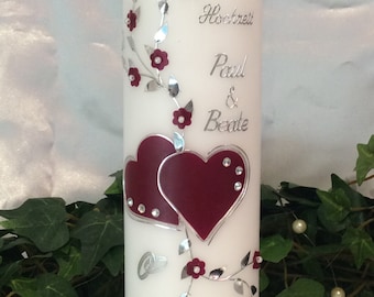 May include: White wedding candle with silver lettering and two burgundy hearts. The candle is decorated with silver vines and burgundy flowers. The text on the candle reads "Zur Hochzeit Paul & Beate 28.06.2023".