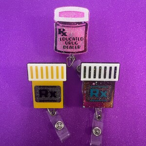 May include: Three prescription-themed badge reels. One pink glitter badge reel with the text "EDUCATED DRUG DEALER". Two additional badge reels with the Rx symbol, one yellow and one with a colorful glitter design.