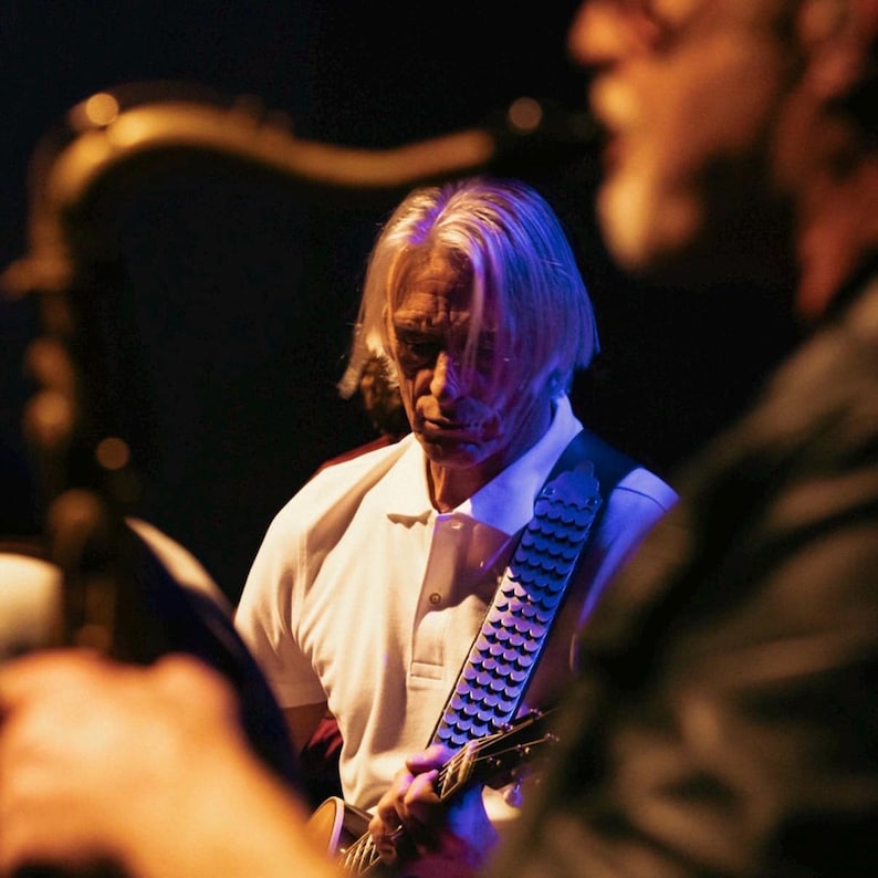 May include: A musician with light hair wears a white polo shirt and plays a guitar with a textured, metallic-looking strap. The scene is dimly lit with blue and warm orange lighting, focusing on the performer's upper body and the instrument.