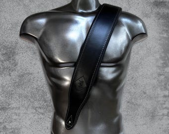 May include: A black guitar strap featuring a wide, padded design with visible light-colored stitching along the edges. The strap includes a diamond-shaped embossed logo patch near the bottom and a reinforced triangular attachment point for a guitar.