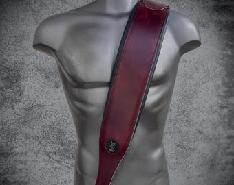 May include: A wide guitar strap featuring a deep burgundy, marbled finish with black leather edges. The strap is displayed on a gray mannequin torso and includes a small, circular black emblem near the bottom end with a white graphic logo.