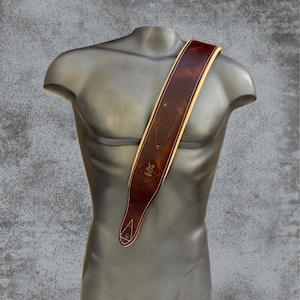 Può includere: Un'ampia tracolla per chitarra in pelle marrone scuro con finitura liscia e lucida e cuciture di colore chiaro lungo i bordi. La tracolla presenta un inserto decorativo in pelle con tre intagli verticali e un piccolo logo in rilievo.