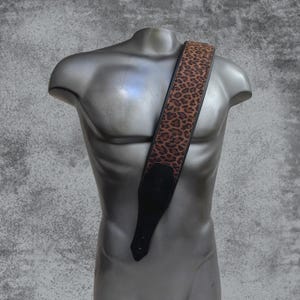 Puede incluir: Una correa de guitarra con un patrón de estampado de leopardo marrón y negro en la sección principal, bordeada por bordes negros. La correa incluye extremos de cuero negro sólido con un diseño de costuras reforzadas y un pequeño logo en relieve.