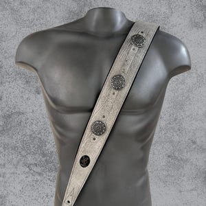 Op de afbeelding: Een lichtgrijze gitaarband met drie zilverkleurige, cirkelvormige versieringen. De band wordt op een mannequin getoond en heeft een textuur en een zwarte onderkant. De band heeft een logo aan de onderkant.