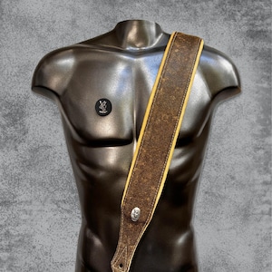 Puede incluir: Una correa de guitarra de cuero marrón con ribete amarillo y hebilla plateada. La correa está desgastada y tiene un aspecto desgastado.