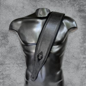 May include: A black leather guitar strap with a diamond-shaped logo featuring a stylized anchor. The strap has a black border and is shown on a mannequin.