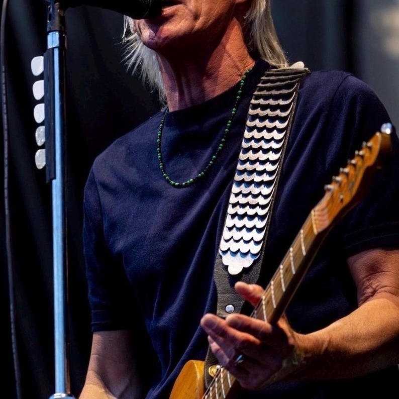 May include: A dark blue t-shirt worn with a guitar strap featuring a silver-toned metallic scale pattern. The strap is attached to a wooden electric guitar. A green beaded necklace is worn around the neck, and white guitar picks are on the mic stand.