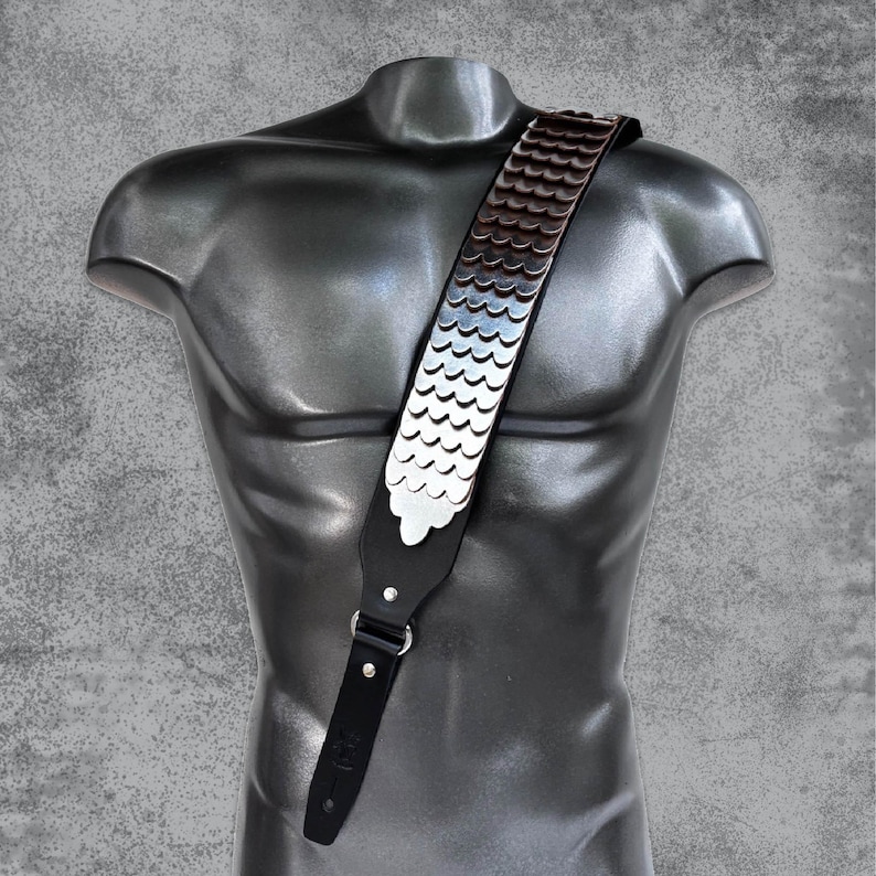 May include: A black leather guitar strap with a silver and brown scale pattern. The strap is adjustable and has a buckle closure.