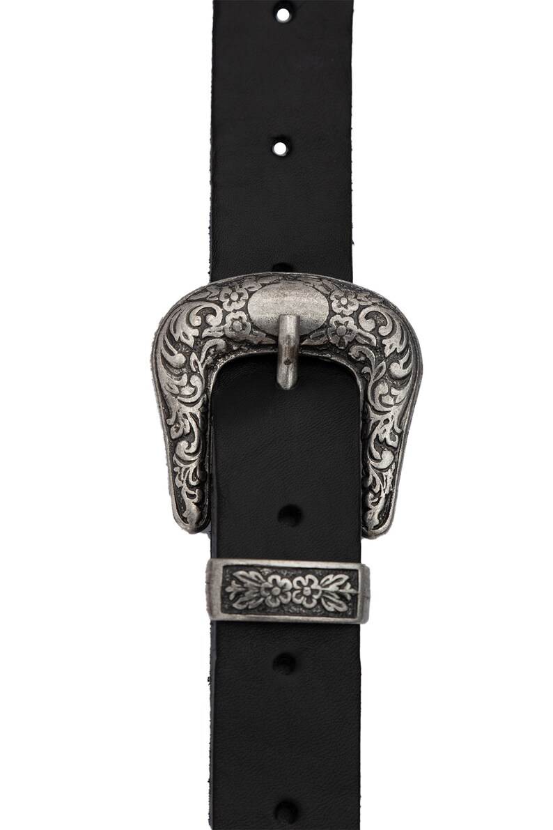 May include: A black leather belt with a silver ornate buckle. The buckle has a floral design and a rectangular piece with a floral design.
