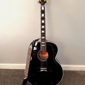 May include: A black acoustic guitar with a glossy finish, featuring a Gibson headstock logo, gold tuning pegs, a dark fretboard with rectangular mother-of-pearl inlays, a round sound hole, and a black guitar strap with a silver metallic pattern.