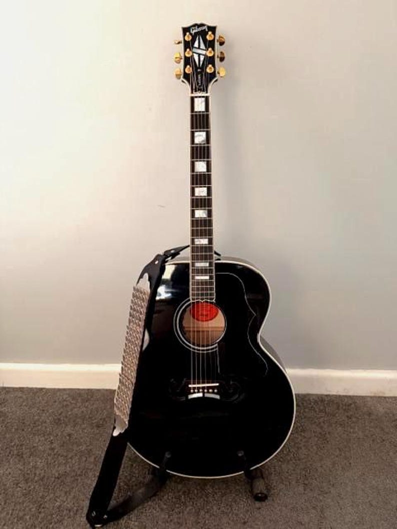 May include: A black acoustic guitar with a glossy finish, featuring a Gibson headstock logo, gold tuning pegs, a dark fretboard with rectangular mother-of-pearl inlays, a round sound hole, and a black guitar strap with a silver metallic pattern.