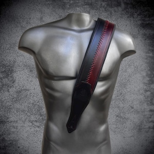 Pu&ograve; includere: Una tracolla per chitarra caratterizzata da un corpo largo in pelle nera con un pannello centrale in pelle color bordeaux intenso. Le due sezioni sono unite da un motivo decorativo a punto croce con filo rosso e terminali in pelle nera.