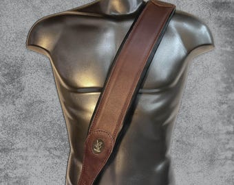 May include: A brown leather guitar strap with a gold metal logo. The strap is shown on a mannequin.