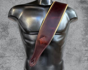 May include: A brown leather guitar strap with a yellow stitched border and a small square logo. The strap is shown on a mannequin.