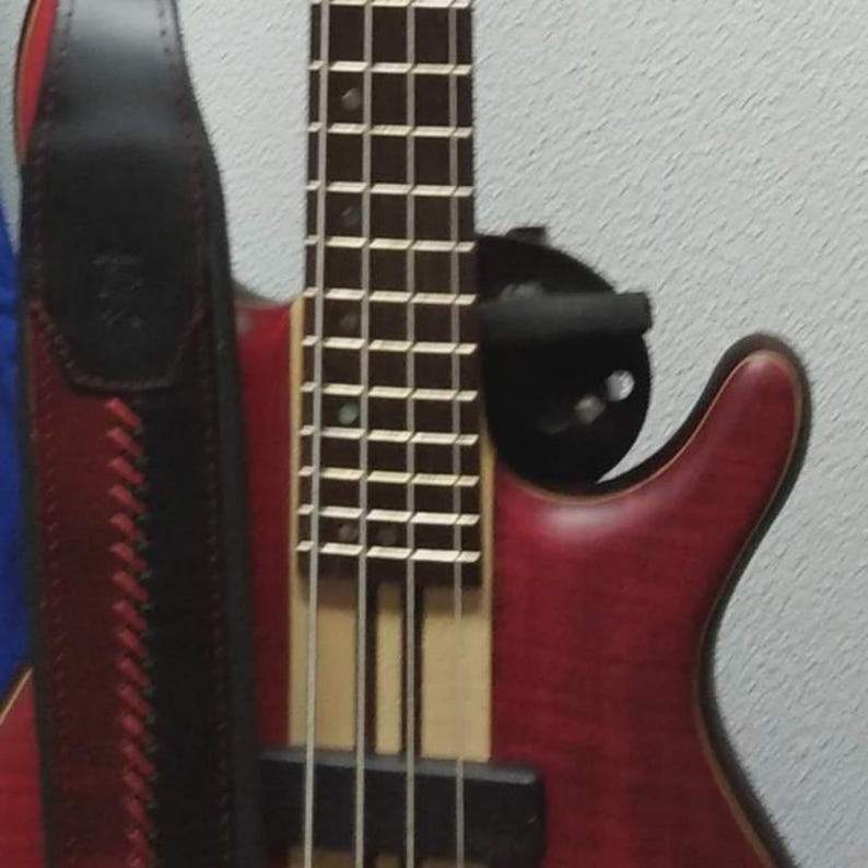 Pu&ograve; includere: Un basso elettrico rosso con una striscia centrale in legno naturale e una tastiera scura, dotato di una tracolla in pelle nera con cuciture rosse. Lo strumento ha una finitura liscia ed &egrave; montato su un supporto da parete nero.