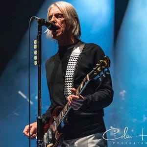 May include: A person with shoulder-length blonde hair wearing a black long-sleeve shirt and a white guitar strap with a repeating circular pattern. The person is playing a dark-colored electric guitar while singing into a black microphone on a stand.