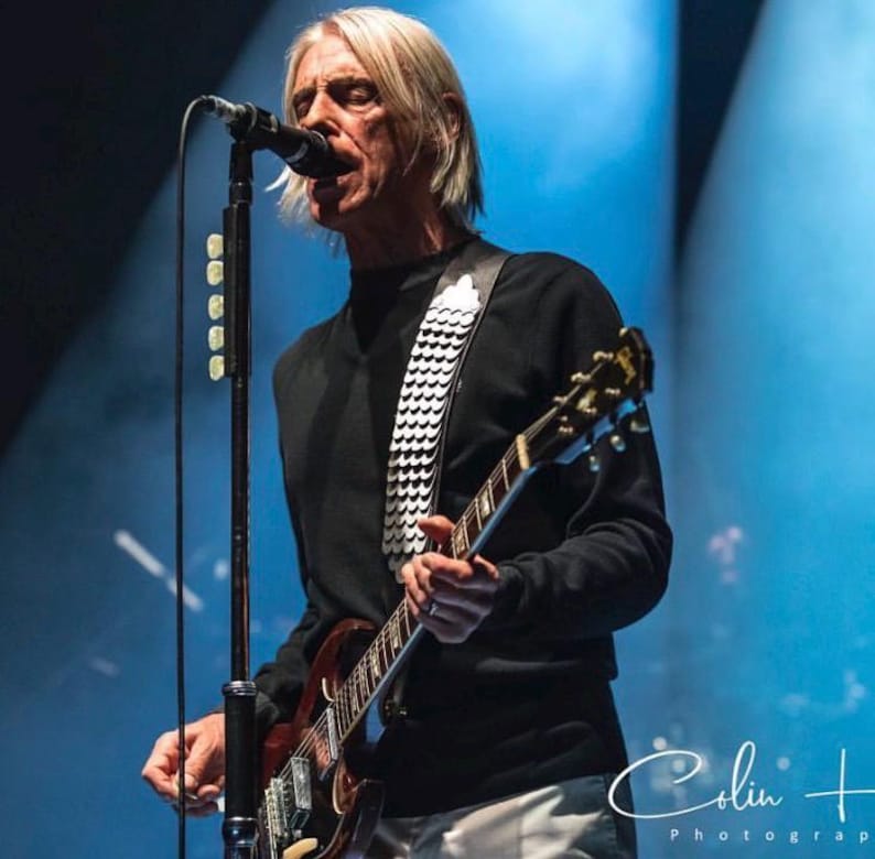 May include: A person with shoulder-length blonde hair wearing a black long-sleeve shirt and a white guitar strap with a repeating circular pattern. The person is playing a dark-colored electric guitar while singing into a black microphone on a stand.