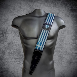 May include: A blue and white guitar strap with a Greek flag design. The strap is made of leather and has a black adjustable strap.