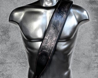 May include: A black leather guitar strap featuring an embossed sugar skull and floral pattern. The strap has a smooth, dark finish with visible stitching along the edges and a reinforced end piece with a pre-cut hole for attachment to a guitar.