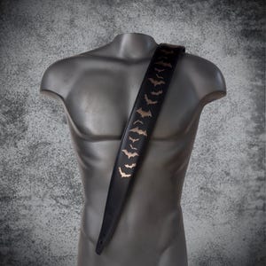 May include: Black guitar strap with a repeating pattern of gold-colored bats. The strap is displayed on a mannequin. The bats are in various sizes and are evenly spaced along the length of the strap. The background is a textured gray.