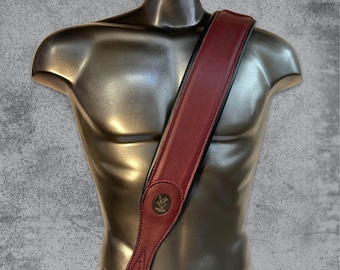 May include: A burgundy leather guitar strap with a brown leather trim and a metal logo. The strap is shown on a mannequin.