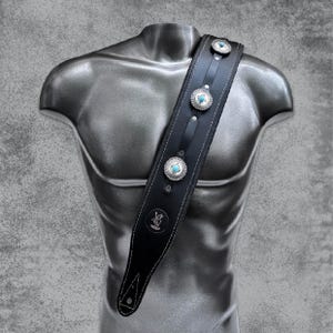 Op de afbeelding: Zwarte leren gitaarband met turquoise en zilveren versieringen. De band heeft witte stiksels en een klein logo aan de voorkant.