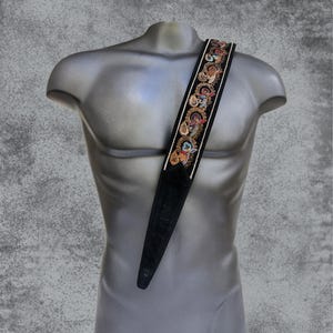 Può includere: Cinturino per chitarra nero con un design ricamato bianco e oro con fiori colorati e volute.
