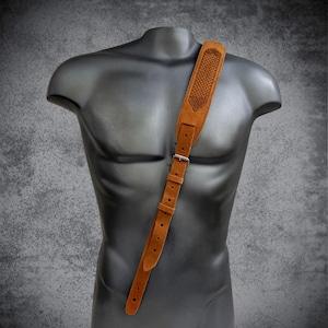 Pode incluir: Uma correia de guitarra de couro castanho com um design tecido na almofada do ombro. A correia é ajustável com uma fivela prateada e apresenta uma série de orifícios para ajuste. A correia é exibida em um manequim preto.