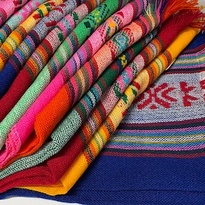 May include: A stack of colorful woven textiles, featuring a variety of vibrant hues including pink, blue, green, and yellow. Each textile has a unique striped or patterned design, with fringed edges. The textiles appear to be approximately 12 inches wide.