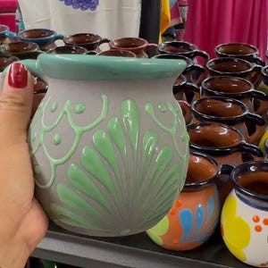 Pode incluir: Uma caneca de cerâmica cinza com uma borda verde e um design floral verde. A caneca tem uma alça e faz parte de uma coleção de canecas artesanais semelhantes com vários designs e cores.