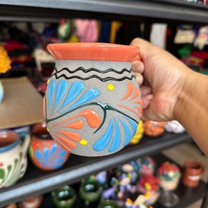 Op de afbeelding: Een handbeschilderde keramische mok met een grijze basis, oranje rand en handvat. De mok is versierd met blauwe en oranje bloemmotieven en een golvende zwarte lijn. Andere kleurrijke aardewerk is zichtbaar op planken op de achtergrond.