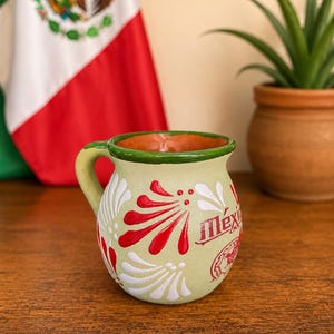 May include: A light green ceramic mug with a rounded body and a handle, featuring hand-painted red and white floral patterns and the word Mexico in red script. The rim is painted in a solid dark green, and the interior is a warm terracotta color.