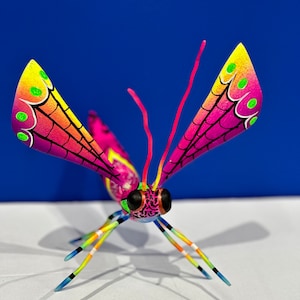 May include: A colourful wooden insect with pink, yellow, green and black wings. The insect has a black and pink head with large green eyes and long, colourful legs.