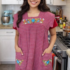 May include: Red and white checked apron with embroidered floral details. The apron features two front pockets and a square neckline. The embroidery includes blue and yellow flowers. The apron is worn in a kitchen.