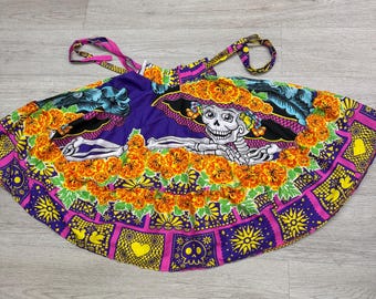 Può includere: Un grembiule svasato con una stampa vivace di uno scheletro che indossa un cappello a tesa larga, circondato da fiori di calendula arancioni brillanti. Il tessuto presenta motivi geometrici viola, rosa e gialli con teschi, cuori e soli.