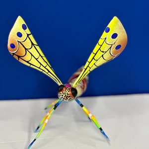 May include: A colourful wooden dragonfly sculpture with yellow wings, blue and orange accents, and black eyes. The dragonfly has six legs and is painted in a vibrant, whimsical style.
