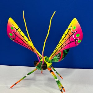 May include: A brightly painted wooden dragonfly sculpture. The wings are shaded from pink to yellow, with green spots and black vein details. The body is green and yellow, with black and orange accents on the legs and antennae. It is set against a blue background.