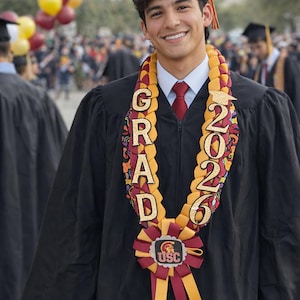 May include: A smiling graduate wearing a black cap and gown, adorned with a graduation lei. The lei features the word "GRAD" and the year "2026" in gold lettering, along with a USC emblem.